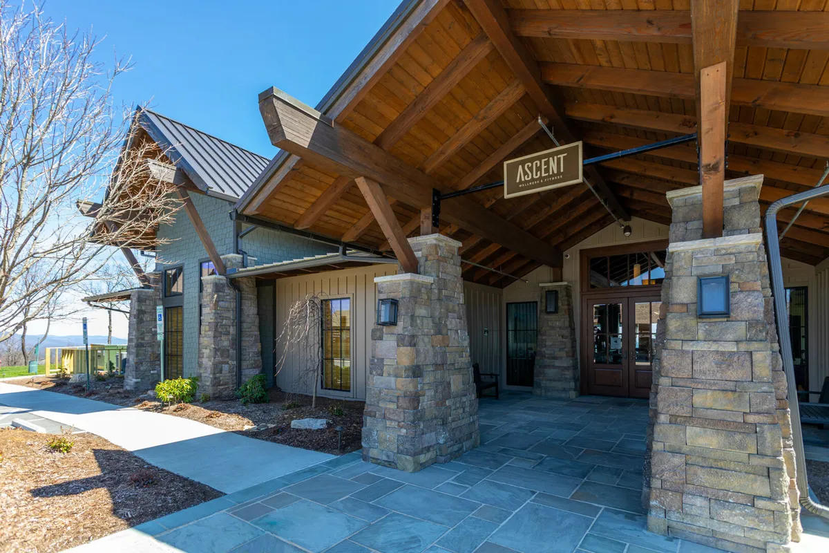 A welcoming exterior shot of the community entrance featuring the prominent 'ASCENT' sign, rustic wooden beams, and sturdy stone pillars. The entrance is framed by natural landscaping, suggesting an inviting and secure environment for guests.