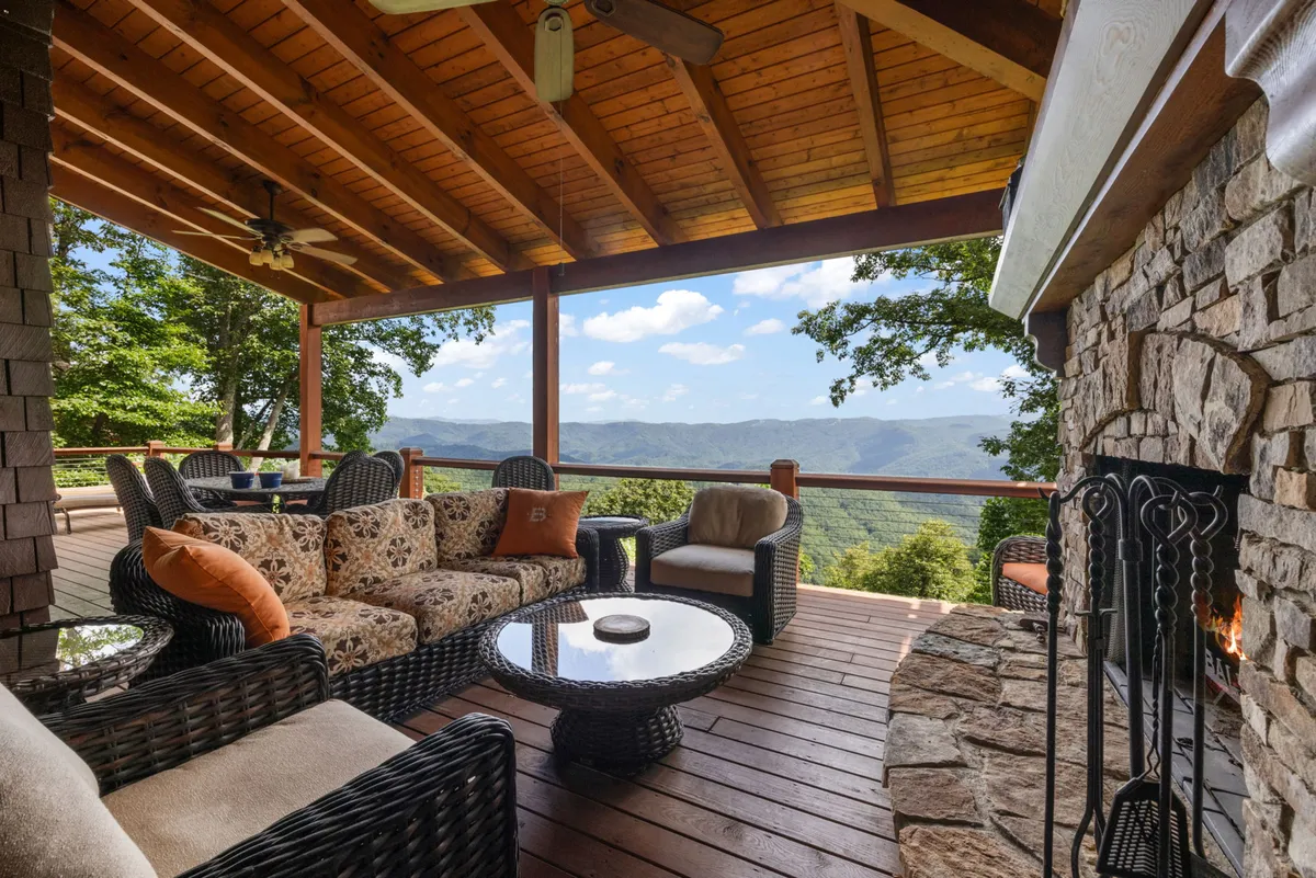 An inviting seating area on the deck overlooks expansive mountain views, centered around a substantial stone fireplace. The setup is ideal for relaxing with family or entertaining guests as the sun sets over the mountains.