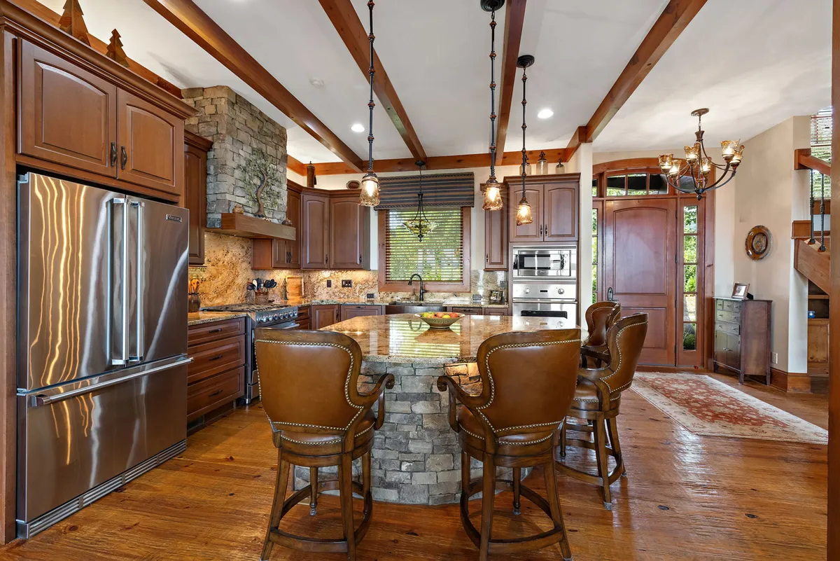 A beautifully designed kitchen with a central island, stone backsplash, pendant lighting, wooden beams, and stainless steel appliances. The layout suggests an open connection to adjacent living and dining areas, with a sense of warmth and communal gathering.