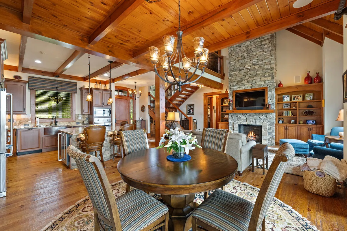 A cozy interior scene featuring a dining table set beneath a chandelier, with a stone wall and fireplace providing a warm backdrop. The space is open and welcoming, ideal for family dining or entertaining guests in a comfortable setting.