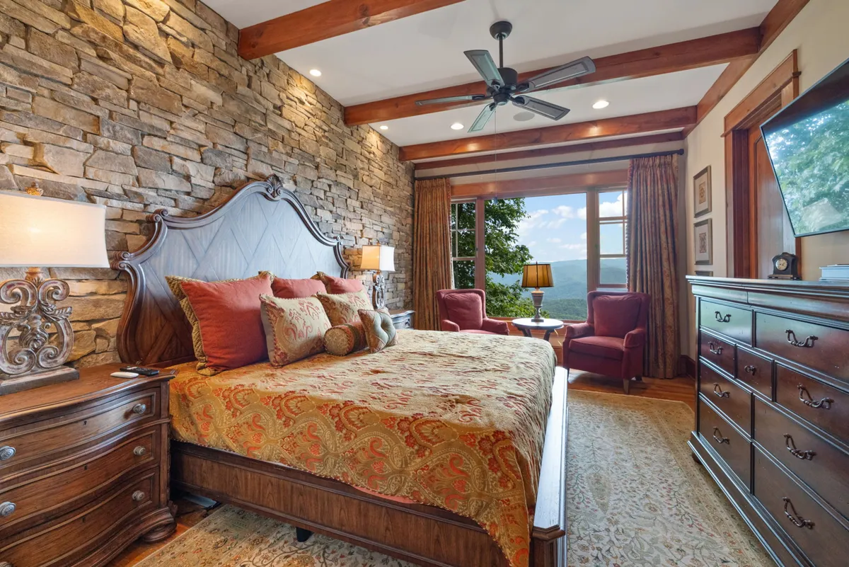 This image features a serene bedroom suite with a prominent stone accent wall behind the bed, plush decorative pillows, armchairs with a lamp, and a large window that offers sweeping views of the mountains. The composition highlights both comfort and the visual connection to the outdoors, capturing the sense of retreat and luxury emphasized in the section.