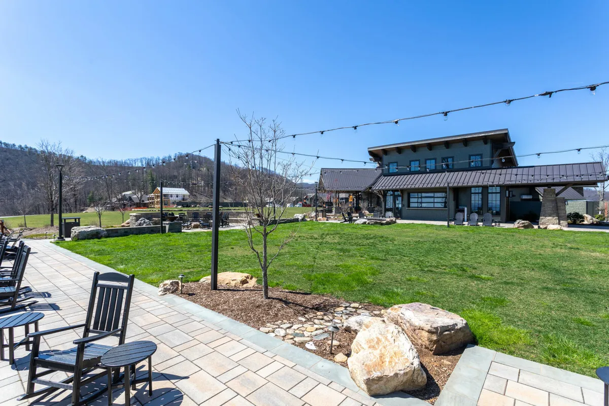 A tranquil exterior scene featuring a modern building set against distinctive rock formations and a well-maintained lawn area. String lights add a festive ambiance, and people are shown relaxing on rocking chairs or gathering for outdoor events, with sunset lighting enhancing the atmosphere.