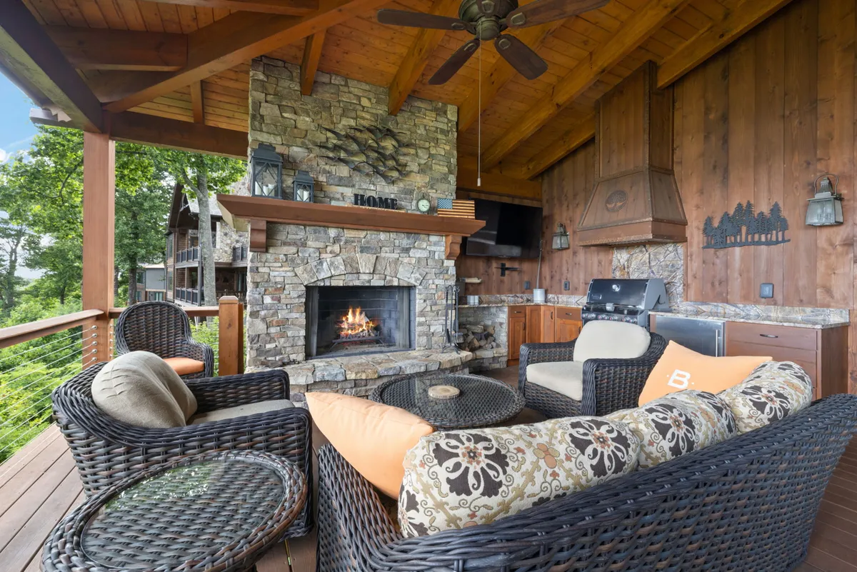 A spacious covered deck features a handsome stone fireplace, a comfortable outdoor seating area, a kitchenette with a built-in grill, and a wooden ceiling. The scene invites guests to gather around the fire, grill meals, and enjoy the mountain air in a sheltered space.