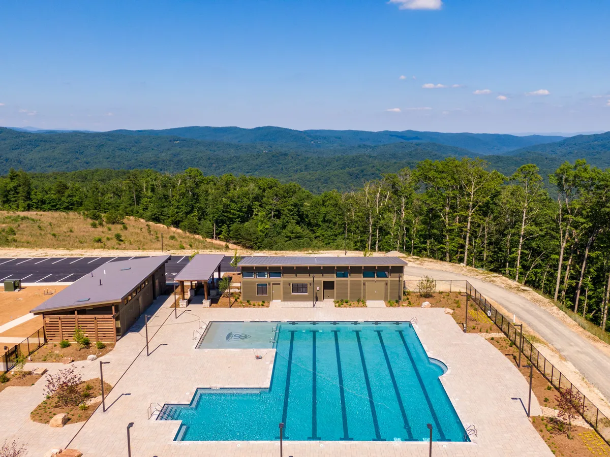 A stunning large swimming pool sits at the base of surrounding mountains, flanked by a modern pool house and landscaped greenery. The tranquil, sunlit setting invites visitors to swim, sunbathe, or simply relax while immersed in the natural beauty of the Blue Ridge.