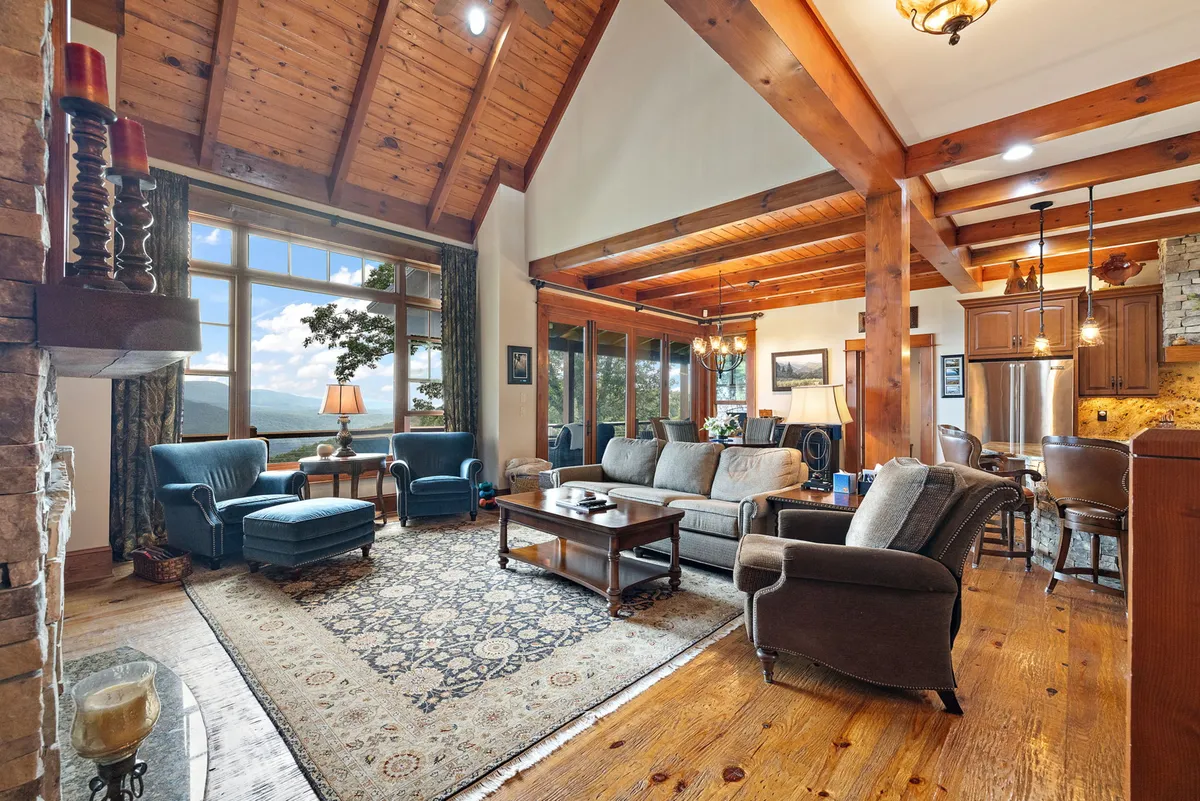 This image showcases a spacious living room with large picture windows framing magnificent mountain views. The room features wooden beams and ceiling, a prominent stone fireplace, and a comfortable seating arrangement—likely plush leather sofas—perfect for gathering and relaxation. The natural light highlights the architectural features while bringing the outdoors in.