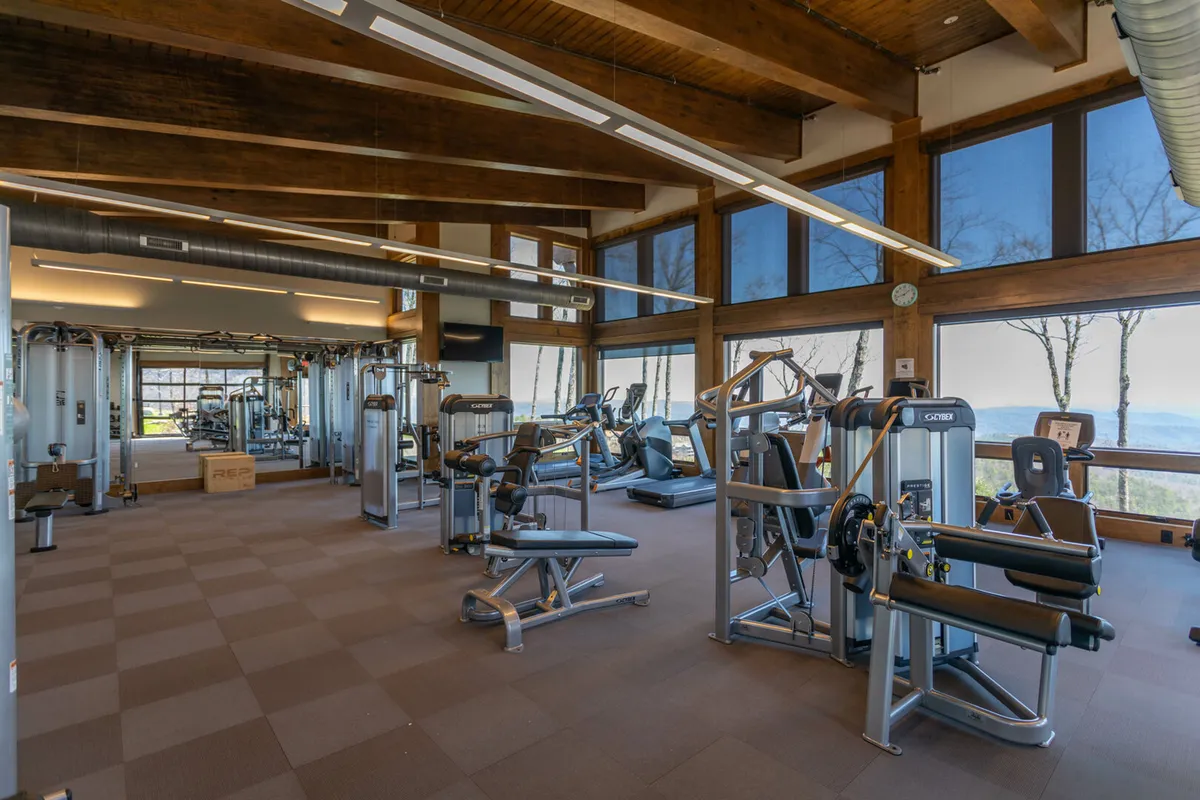 A spacious gym interior with a variety of fitness equipment, large windows flooding the room with natural light, and views of the surrounding landscape. The room is active, with people engaged in workouts and group fitness activities.