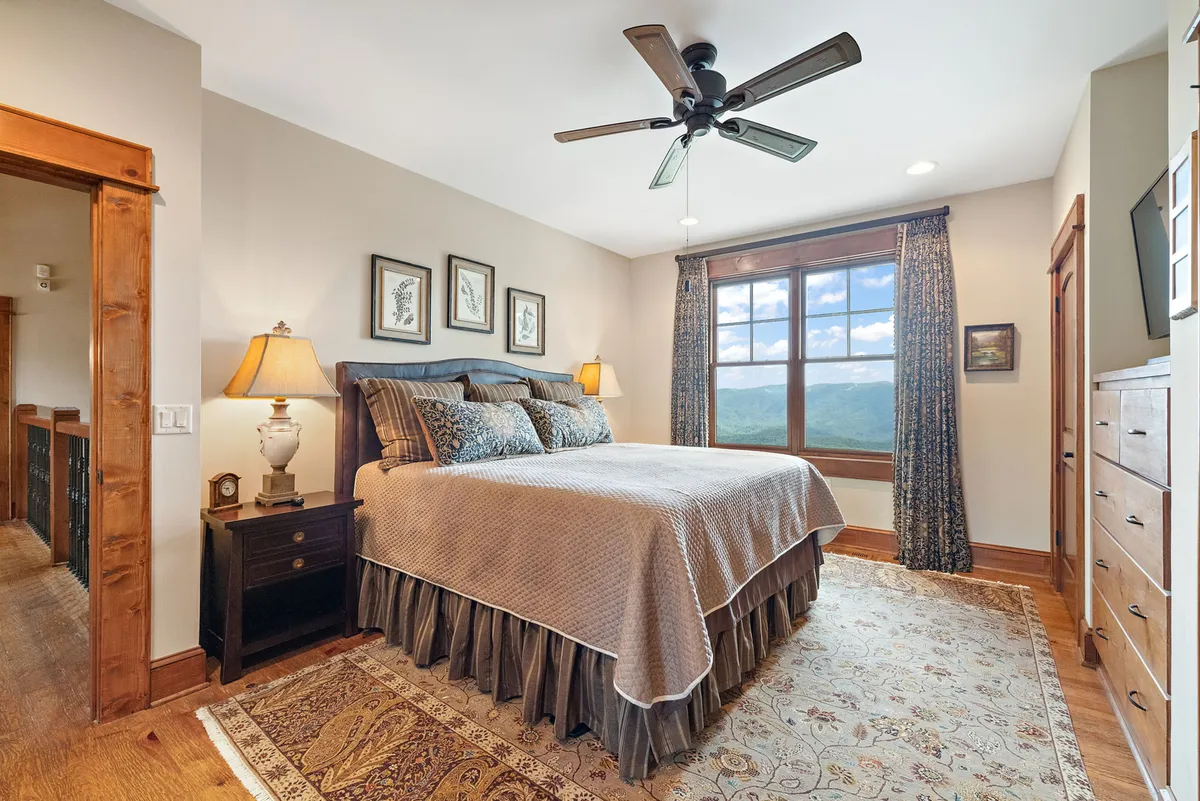 This image shows a tranquil bedroom with a bed adorned with decorative pillows, a nightstand and lamp, and a large window that frames a scenic mountain view. The simplicity and comfort of the room evoke a restful atmosphere, ideal for a peaceful morning start.