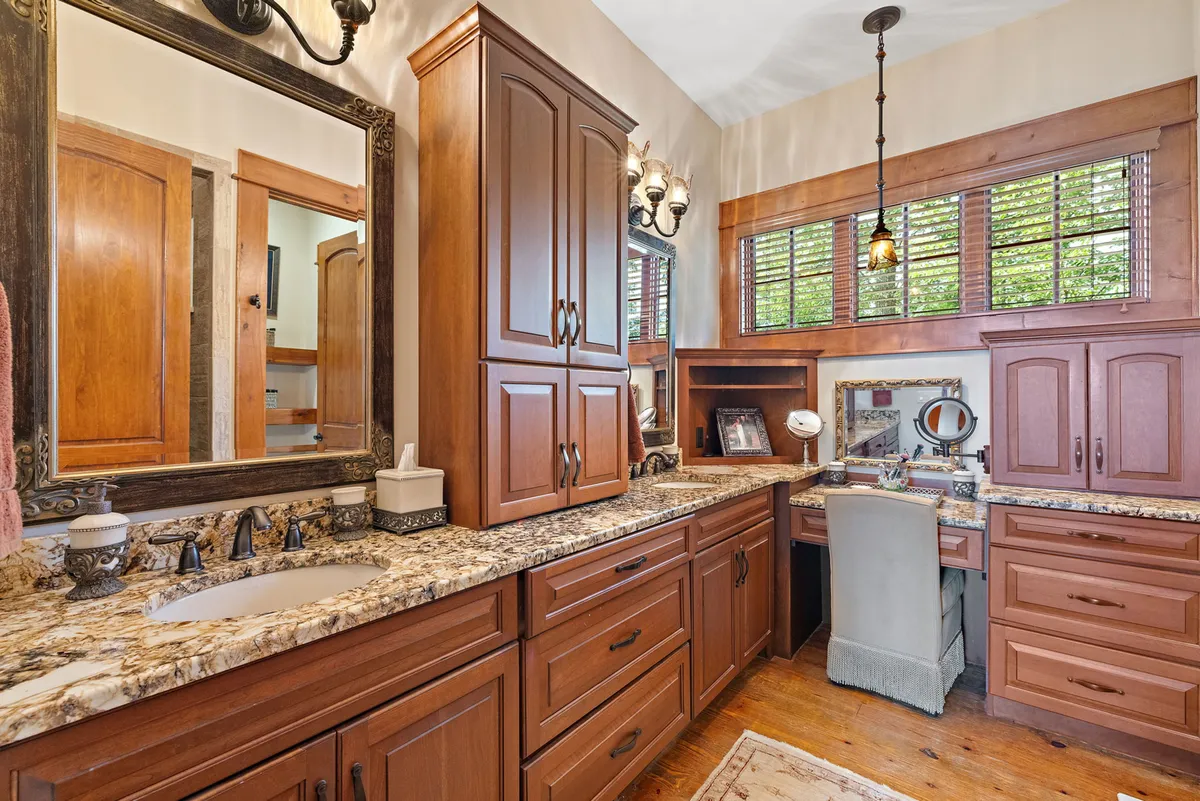 A calm, well-lit interior space featuring a vanity with granite countertop, dark wood cabinetry, and, notably, a desk area with a chair set by a window. The scene feels organized, making it suitable for both working and quiet reflection.