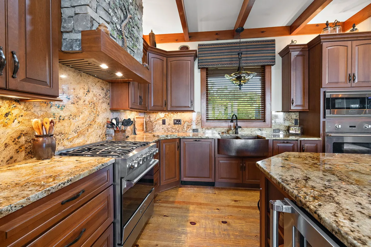 A view of the kitchen area with granite countertops, a stone feature wall, wooden beams, and a large window offering a scenic vista. The kitchen flows naturally into the living and dining space, promoting both entertaining and everyday family life.