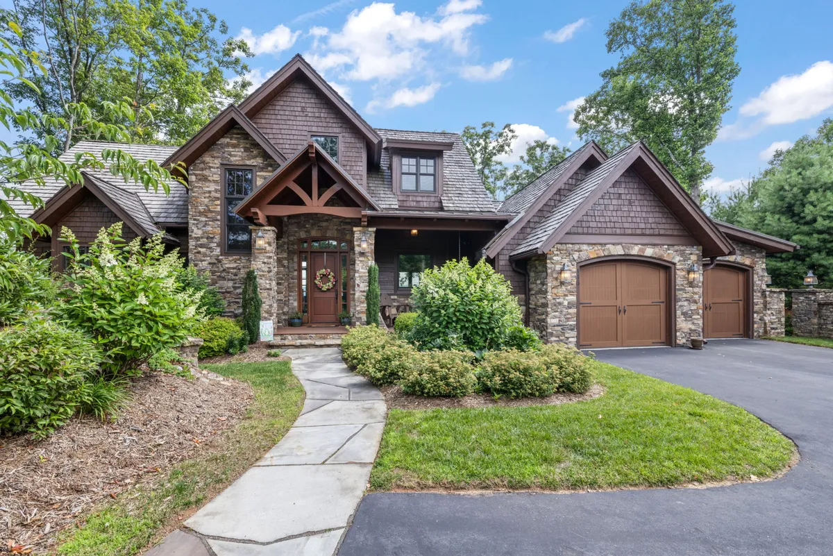 The image showcases the warm stone facade and striking wooden beams of the home, framed by a lush, landscaped garden and a festive wreath on the front door. The inviting porch hints at relaxing mornings or cozy evenings overlooking the mountains, while the surrounding greenery suggests ample space for outdoor gatherings and family fun.